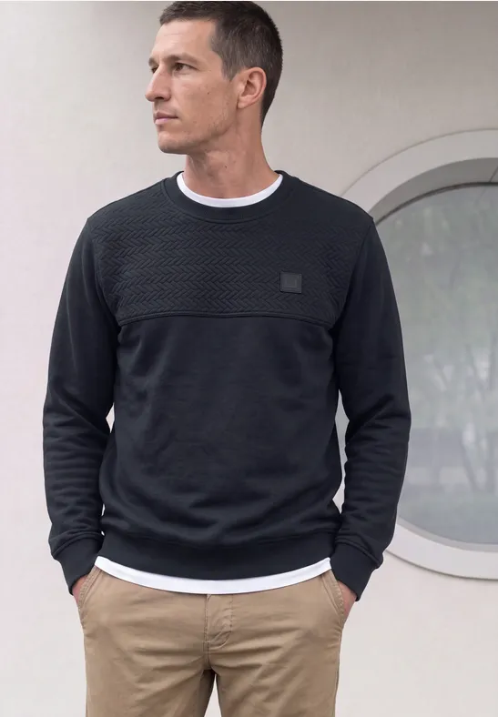 Un homme porte un pull noir, un t-shirt blanc et un pantalon beige, mains dans les poches.