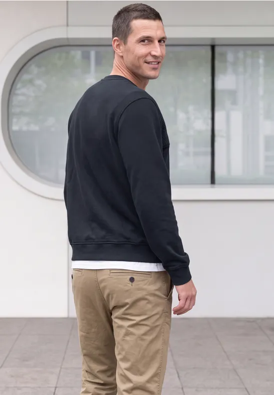 Homme en pull noir et pantalon beige regarde par-dessus l'épaule devant un bâtiment moderne et lumineux.