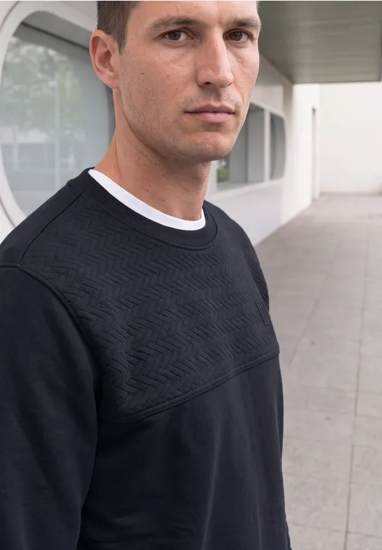 Un homme en pull noir à motif géométrique pose devant un bâtiment moderne.