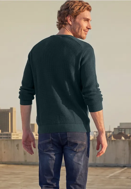Ein Mann in grünem Pullover und Jeans steht mit dem Rücken zur Kamera auf einem Dach.