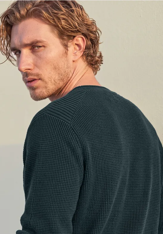 Ein junger Mann mit lockigem blondem Haar im dunklen Pullover blickt über die Schulter zur Seite.