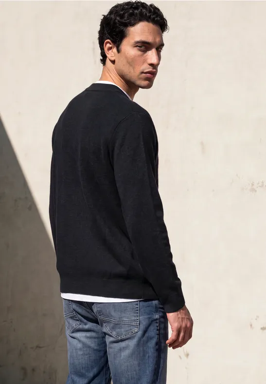 Ein junger Mann steht vor einer hellen Wand und trägt einen schwarzen Pullover und blaue Jeans.