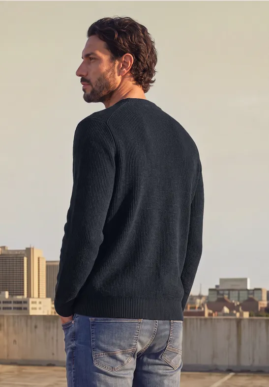 Ein Mann mit dunkelblauem Pullover und Jeans steht auf einem Dach und blickt auf die Stadt.