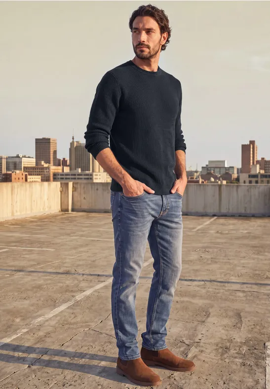Ein Mann in dunklem Pullover und Jeans steht auf einem Parkdeckdach mit Stadthintergrund.