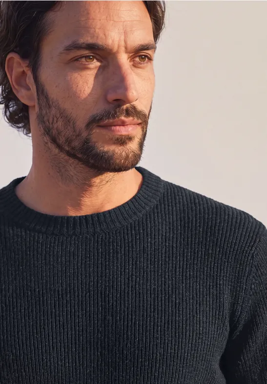 Nahaufnahme eines nachdenklich wirkenden Mannes mit dunklem Pullover, Bart und leicht gewelltem Haar.