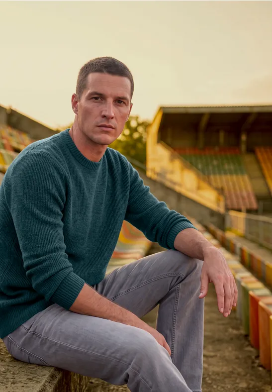 Ein Mann sitzt in einem alten Stadion auf rot-gelben Holztribünen, trägt grünen Pullover und graue Hose.