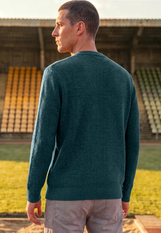 Mann im grünen Pullover steht mit dem Rücken zur Kamera vor leeren Tribünen im Freien.