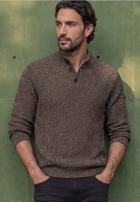 Ein Mann mit dunklen Haaren trägt einen braunen Strickpullover und steht vor einer grünen Wand.