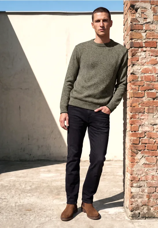 Ein Mann lehnt im Sonnenlicht an einer Backsteinwand, trägt grünen Pullover, dunkle Hose und braune Boots.