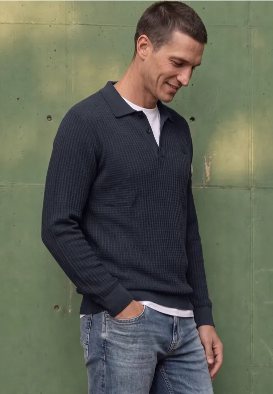 Mann im dunkelblauen Strick-Poloshirt mit weißem Kragen und Jeans vor grüner Wand, lächelnd.