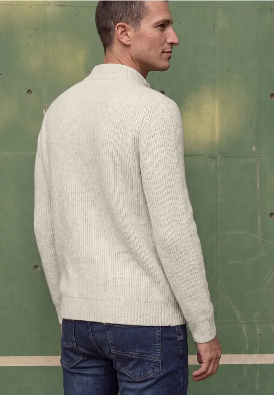 Un homme en pull clair et jean, vu de dos, se tient devant un mur vert.
