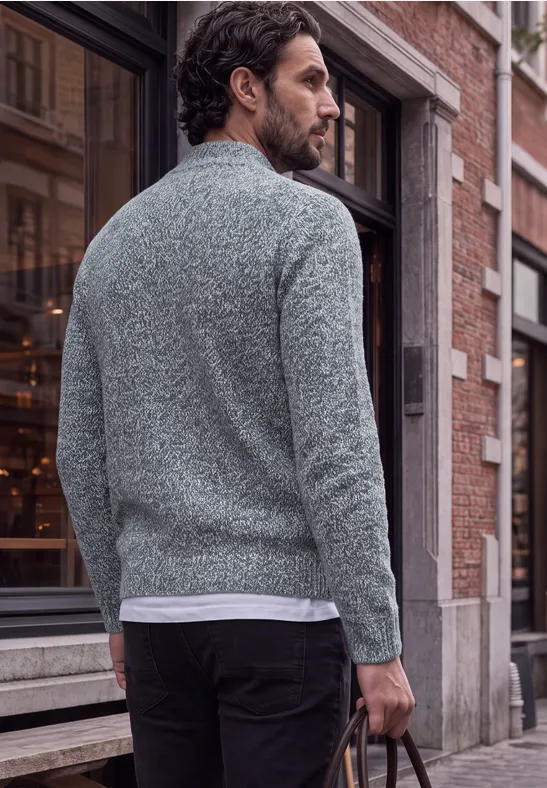 Man in grijze trui en zwarte broek met leren tas in de hand, loopt door de stad.