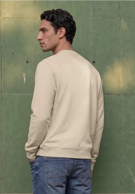 Ein Mann steht seitlich vor einer grünen Wand, trägt beige Sweatshirt und blaue Jeans.
