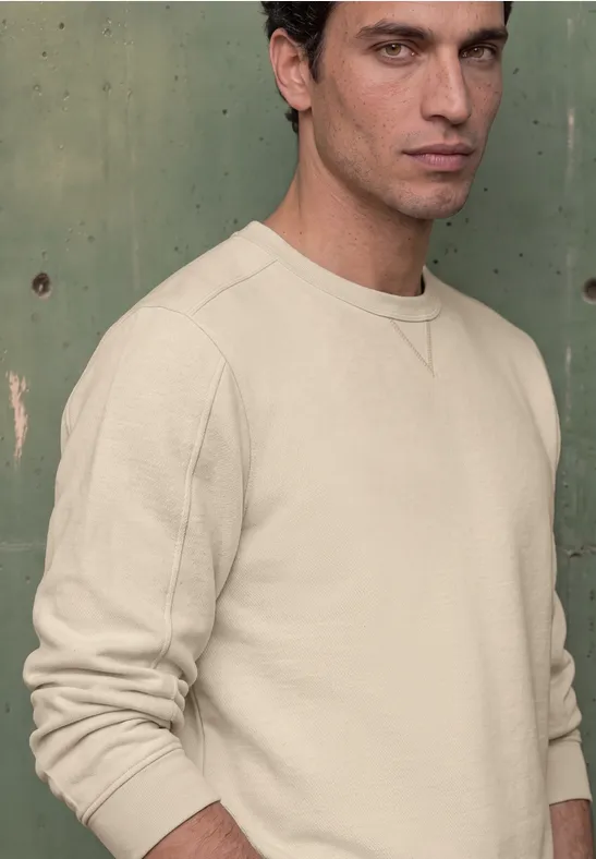 Junger Mann mit dunkelbraunen Haaren in beigem Sweatshirt steht vor einer grünen Wand.