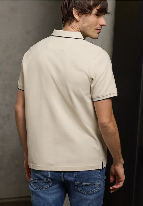 Een man met een beige poloshirt en blauwe spijkerbroek, gezien van achteren voor een lichte muur.