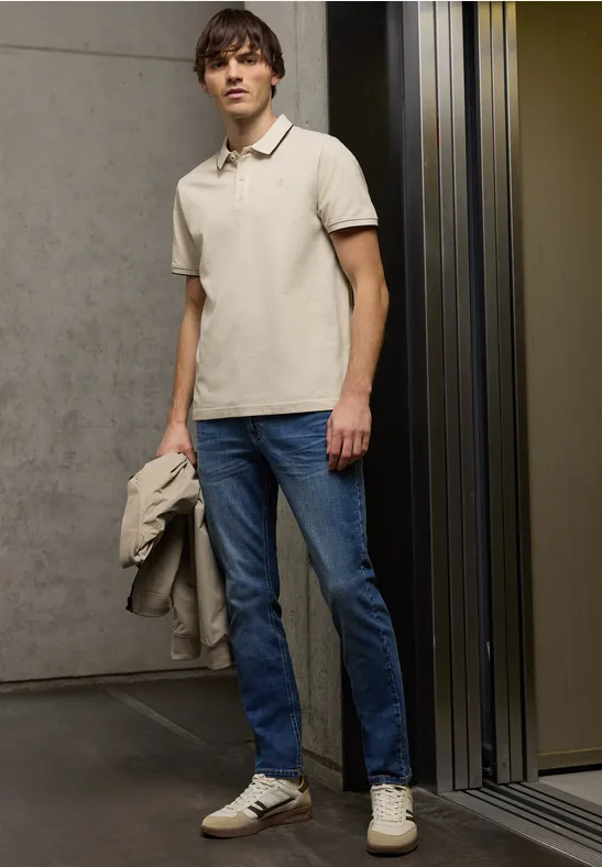 Man in beige polo, spijkerbroek en sneakers, houdt jas vast, staat voor een lift in een moderne gang.