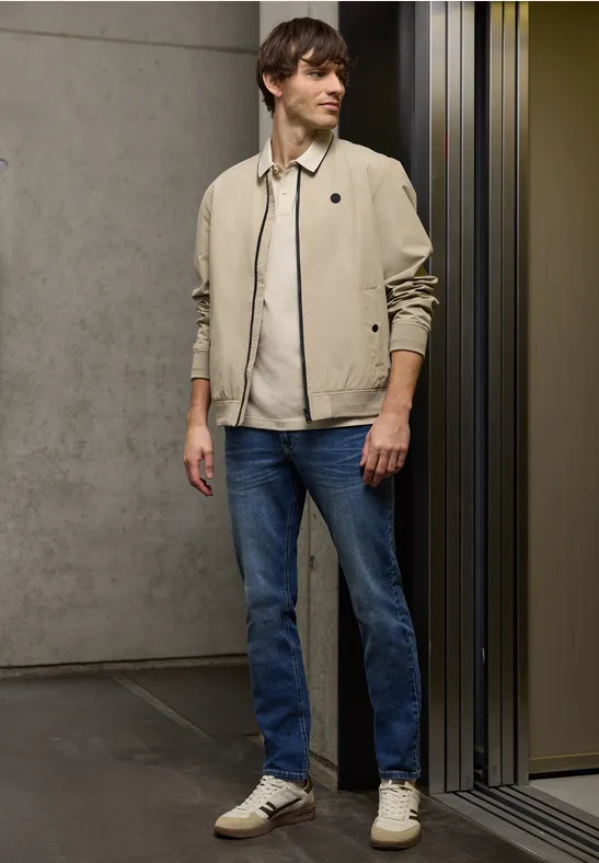 Een man in een lichte jas, jeans en sneakers staat in een moderne gang voor een betonnen wand en glazen deur.