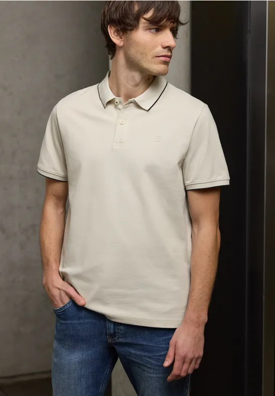 Een man draagt een lichtkleurig poloshirt en blauwe spijkerbroek, staand voor een grijze muur.