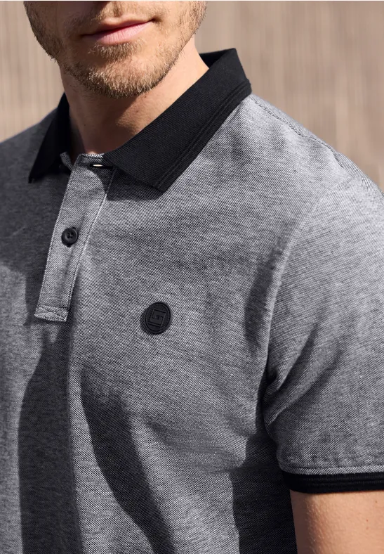 Mann trägt graues Poloshirt mit schwarzem Kragen und Ärmelsäumen, mit Logo auf der Brust.