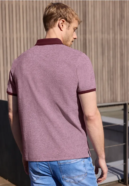 Ein Mann steht mit dem Rücken zur Kamera, trägt ein violettes Polohemd und blaue Jeans vor einer Holzfassade.