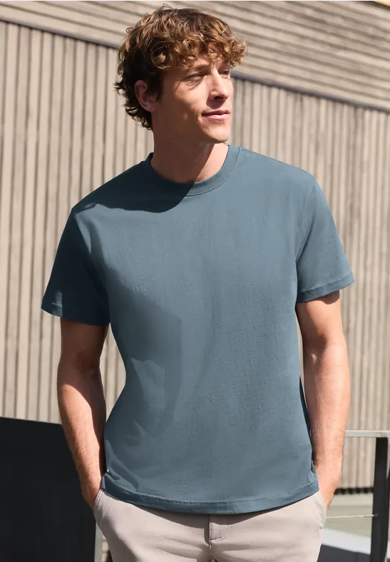 Een man staat ontspannen voor een gevel en draagt een eenvoudig blauw T-shirt.