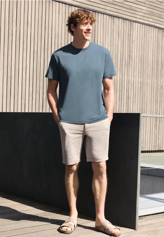 Een man draagt een blauw t-shirt, beige short en sandalen, staat ontspannen voor een houten gevel.