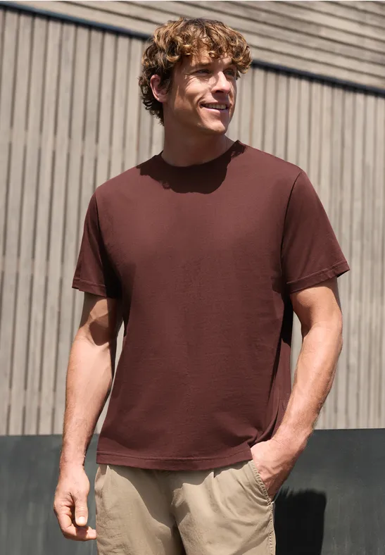 Een man met een bruin T-shirt en beige broek staat voor een houten wand, handen in de zakken.
