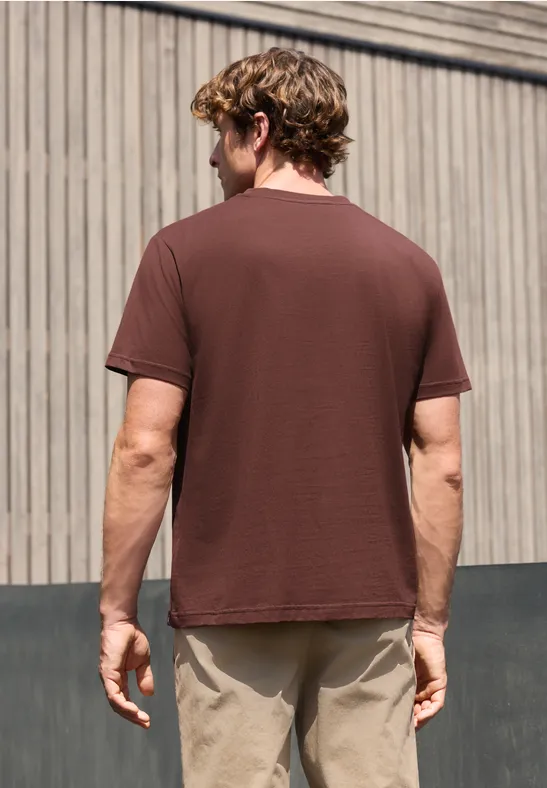 Een man van achteren tegen een houten gevel, bruine t-shirt en beige broek