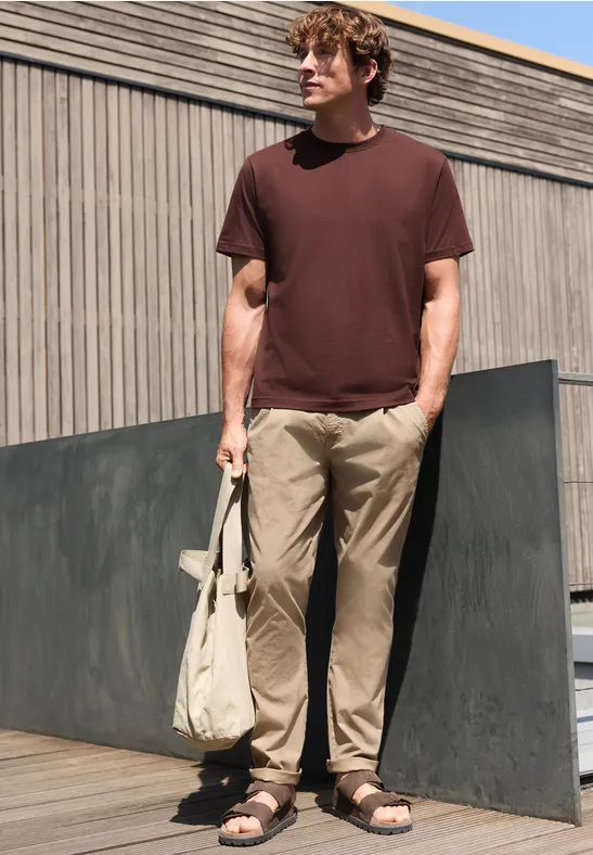 Een man staat buiten voor een houten gevel, draagt een bruin t-shirt, lichte broek en sandalen, en houdt een tas vast.