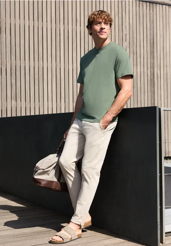 Een man leunt tegen een muur, draagt een groen T-shirt, lichte broek, sandalen en heeft een reistas.