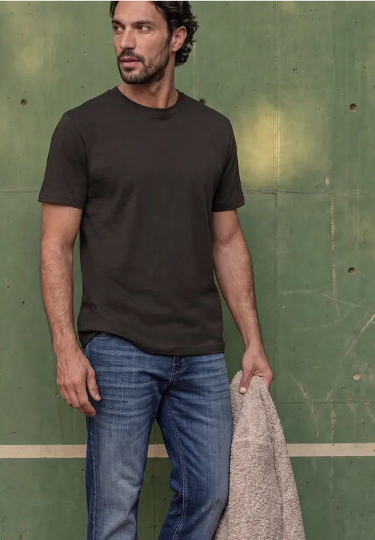 Mann vor grünem Hintergrund trägt dunkles T-Shirt, Jeans und hält eine beige Jacke.
