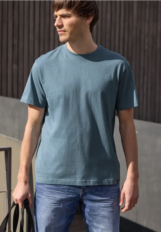 Ein Mann steht vor einer Holzwand, trägt ein blaues T-Shirt, Jeans und hält eine Tasche in der Hand.