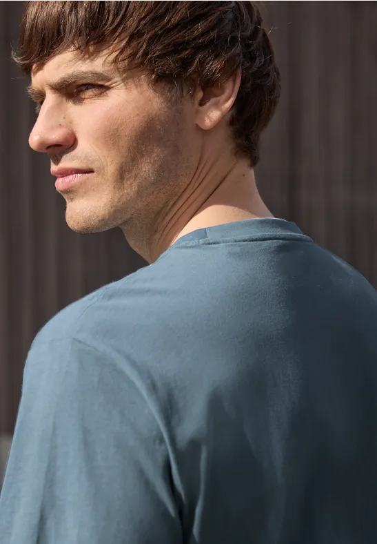 Ein junger Mann steht mit dem Rücken zur Kamera und schaut über seine Schulter, trägt ein blaues T-Shirt.