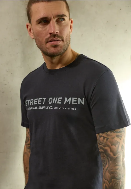 Mann mit dunkelblauem T-Shirt "STREET ONE MEN" und tätowierten Armen vor heller Wand.