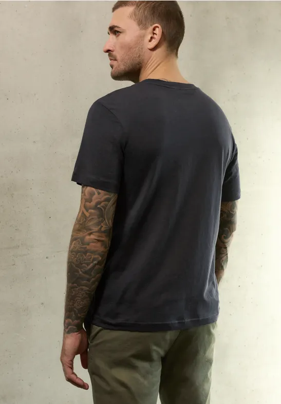 Mann von hinten mit dunkelgrauem T-Shirt, olivgrünen Hosen und tätowierten Armen vor neutralem Hintergrund.