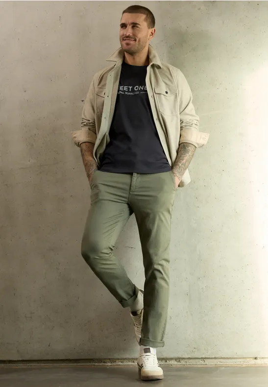 Ein Mann lehnt lässig an einer Betonwand, trägt beige Jacke, dunkelgraues T-Shirt, olivgrüne Hose und weiße Sneaker.