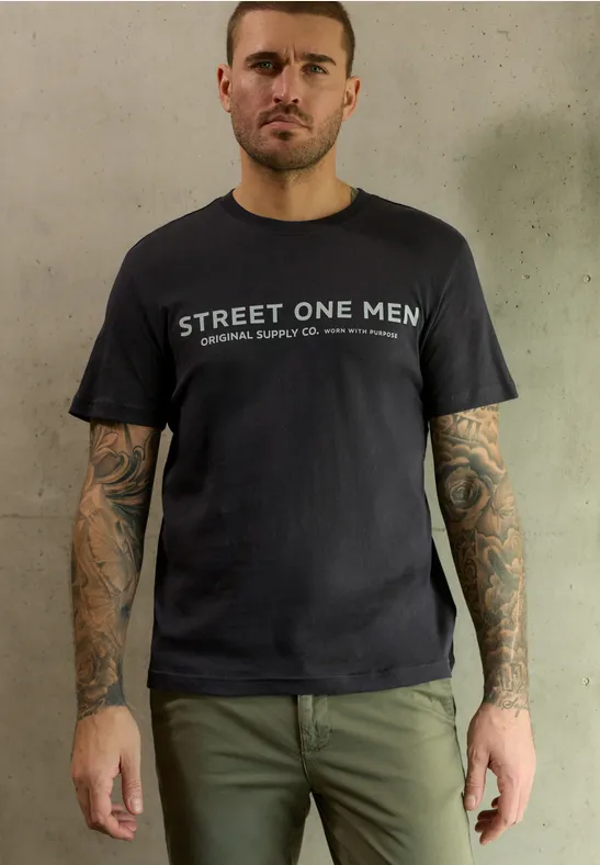 Mann in schwarzem T-Shirt mit STREET ONE MEN-Aufdruck, grüner Hose und tätowierten Armen.