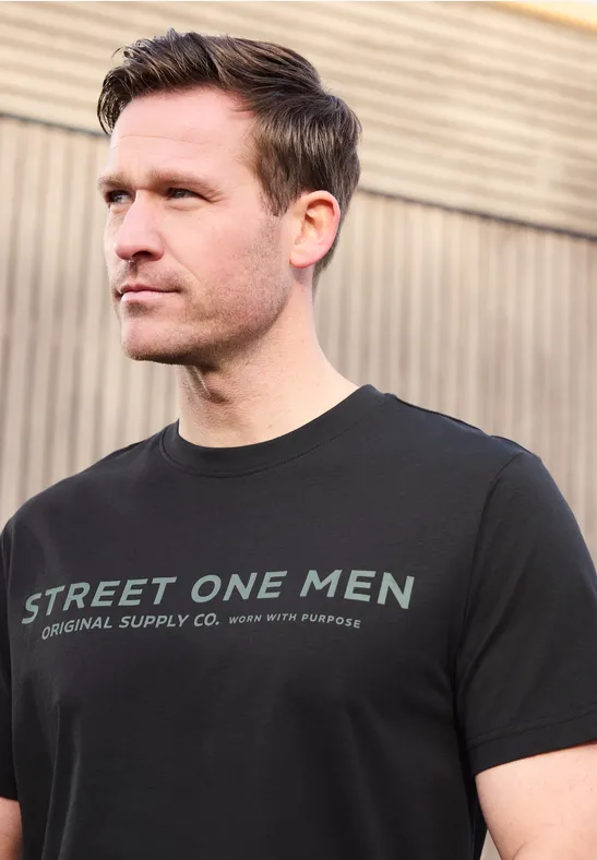 Ein Mann steht vor einer Holzwand und trägt ein schwarzes T-Shirt mit der Aufschrift 'Street One Men'.