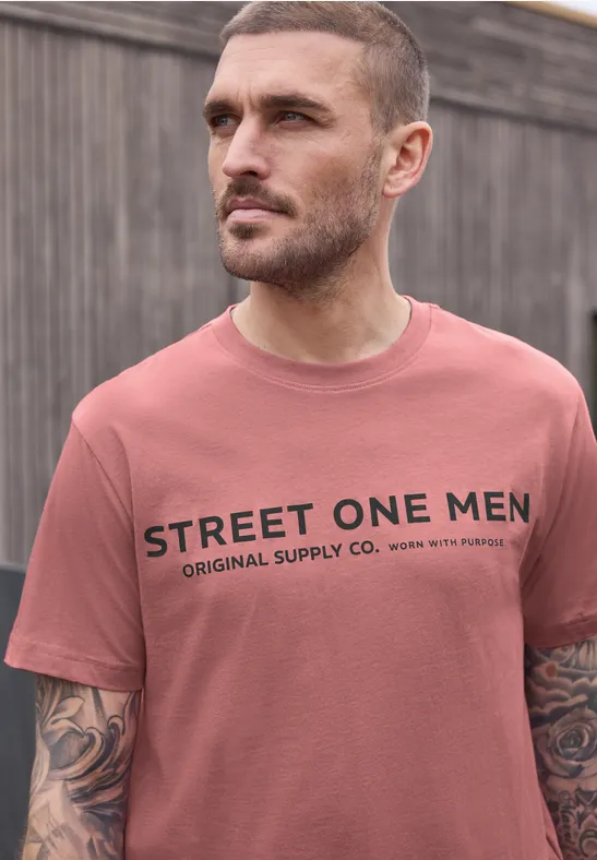 Ein Mann steht vor einem Holzzaun und trägt ein rosafarbenes T-Shirt mit dem Aufdruck „STREET ONE MEN“.