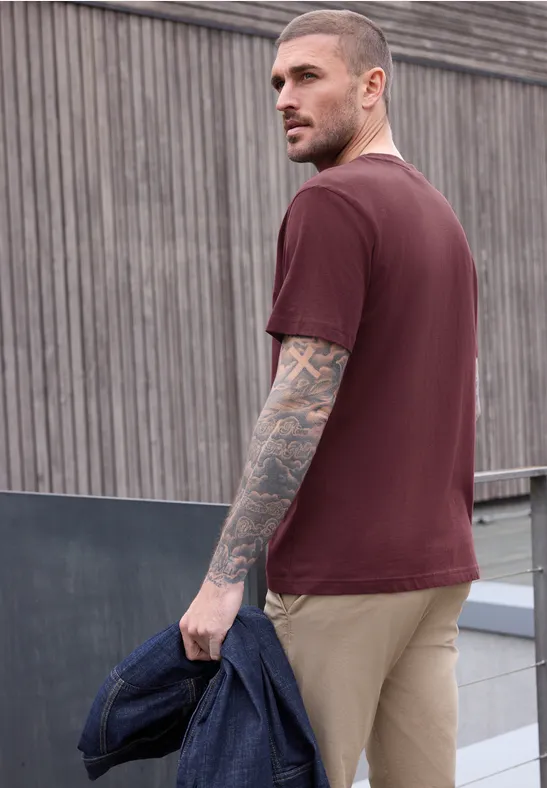 Seitlich abgebildeter Mann mit Tätowierungen, burgunderrotem T-Shirt, khakifarbener Hose und Jacke in der Hand.