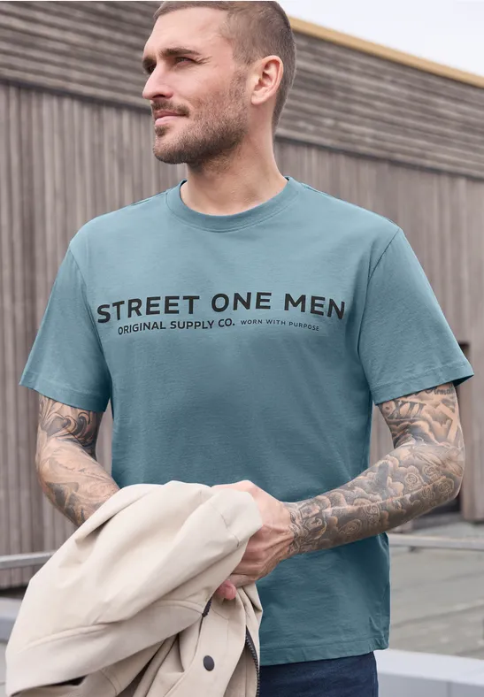 Un jeune homme porte un t-shirt turquoise 'STREET ONE MEN' et tient une veste beige.