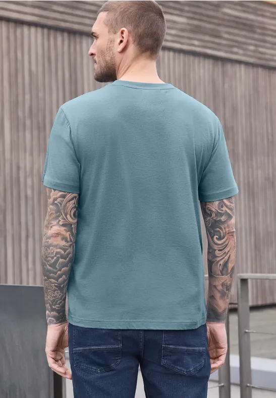 Homme tatoué, vu de dos, en t-shirt bleu et jean, devant un mur en bois.