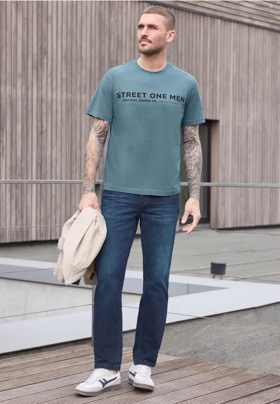 Homme en T-shirt bleu-gris, jeans et baskets, devant un mur en bois, tenant une veste à la main.