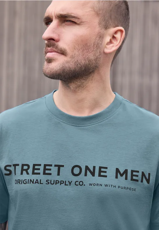 Un homme au visage flouté devant un mur de bois porte un T-shirt turquoise avec l'inscription 'STREET ONE MEN'.