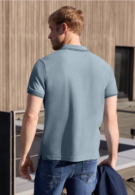 Ein Mann steht von hinten in einem hellblauen Poloshirt und Jeans, hält eine Jacke vor einer Holzfassade.