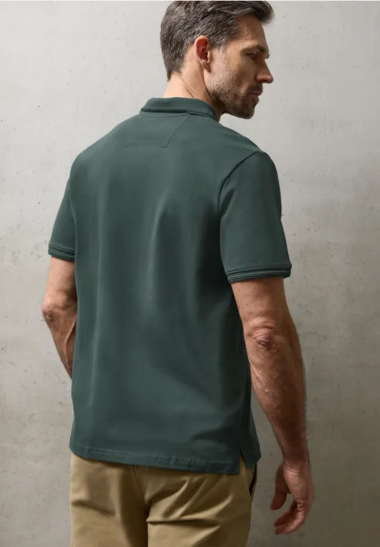 Ein Mann steht vor neutralem Hintergrund, trägt ein dunkelgrünes Poloshirt und eine beige Hose, Rückansicht.
