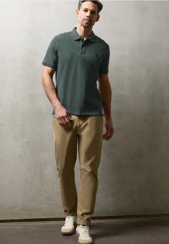 Mann in grünem Poloshirt, beigen Chinos und weißen Sneakers, hält braunes Lederetui, vor neutralem Hintergrund.