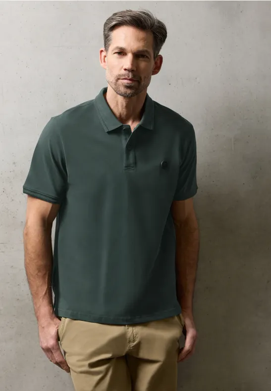 Ein Mann trägt ein dunkelgrünes Poloshirt und beige Hosen, steht vor einer hellgrauen Wand.