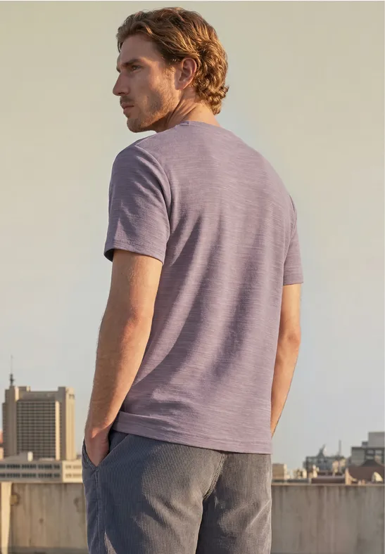 Ein Mann steht auf einem Dach, trägt ein lila T-Shirt und graue Hose und blickt über die Stadt.