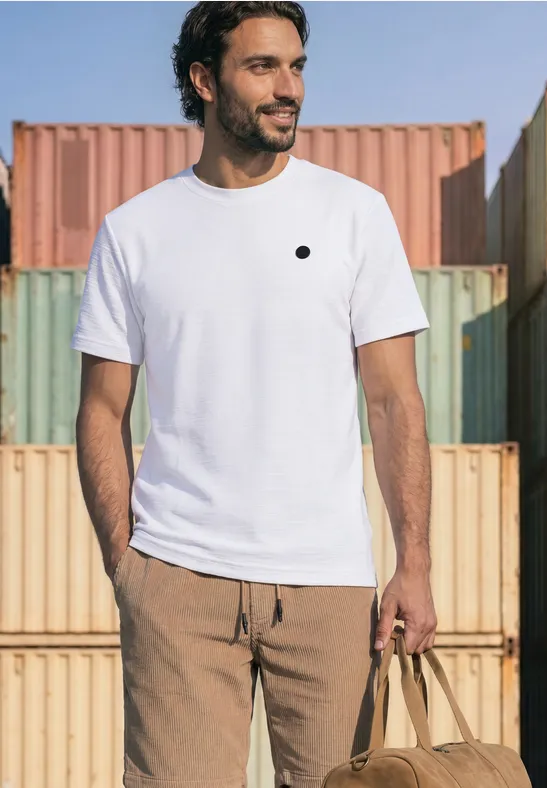 Een man in een wit T-shirt en beige broek poseert met een tas voor kleurrijke containers.